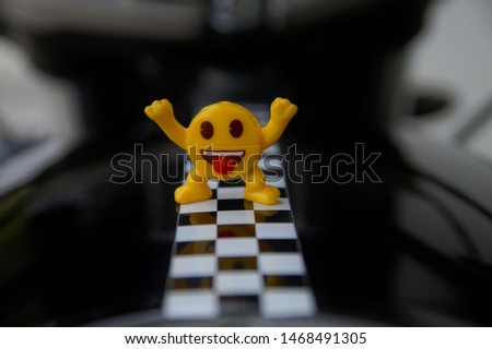 Little yellow smiley man is on motorcycle. winner emoticon smiley jokingly stuck out its tongue and is raising the hands up on racing line. race track with starting or end line, free space for text. 