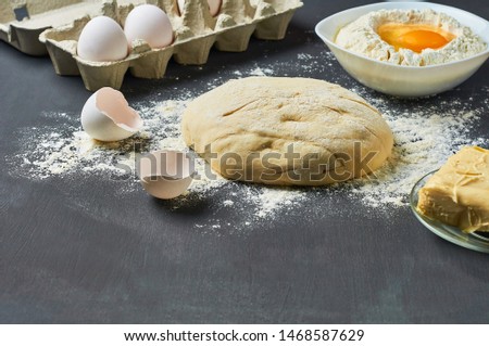 Dough with flour near broken egg with yolk in bowl and other utensil, ingredients lies on dark concrete table. Baking process. Copy space