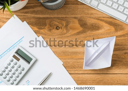Wooden office desk with white origami boat. Professional invest management and consulting. Flat lay office workplace with business reports, computer keyboard and calculator. Individual success