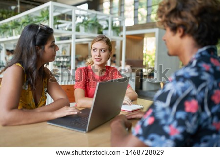 Candid lifestyle shot of multi-ethnic millennial colleague group discussing together on laptop computer at a table in bright modern cafe