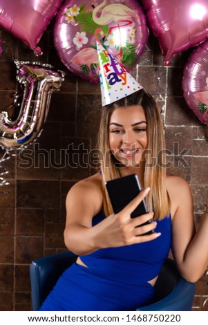 Girl taking a selfie and celebrating a birthday with a cap on head and decorative wall in the background in the restaurant. Birthday celebration concept. Close up, selective focus