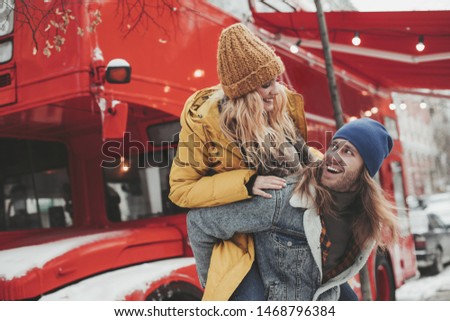 Love is in the air. Waist up portrait of cheerful pretty woman on back of her beloved hipster man leaning on red double-decker bus