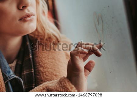 Love is in the air. Close up low face portrait of thoughtful fair haired young girl drawing heart shape on steamy window while sitting in transport