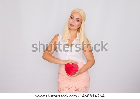 Portrait of a young blonde woman in light-colored clothes. Holding a heart shaped gift box. Posing on a white background.