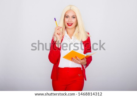 Attractive blonde haired woman wearing red jacket and pants standing isolated over white background, holding orange book and pen in her hands