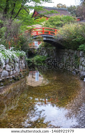 Portrait photo of the shrine grounds in Shimogamo Shrine in Kyoto City with a small river stream leading to a small arched bridge, painted in orange, in the background.