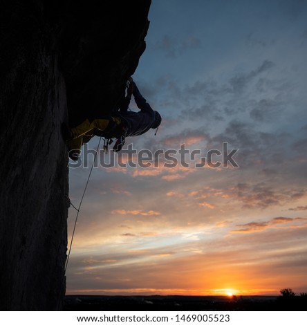 Back low angle view of male climber on rocky wall at sunset conquering the top of steep cliff. Evening sky with sunlight on horizon on background. Active outdoors extreme lifestyle concept. Copy space