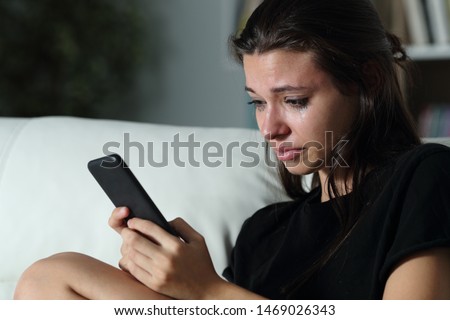 Sad teenage girl checking smart phone cyber bullying messages sitting on a couch in the dark at home Royalty-Free Stock Photo #1469026343