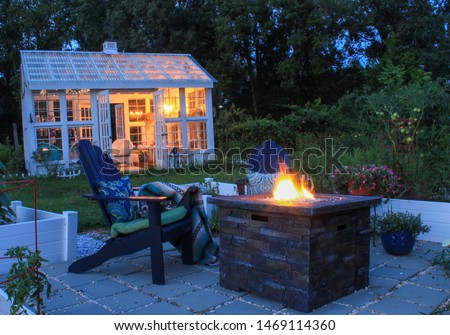 Beautiful Garden Scene at Dusk with Firepit, Andirondack Chairs and Greenhouse with lit chandelier Royalty-Free Stock Photo #1469114360