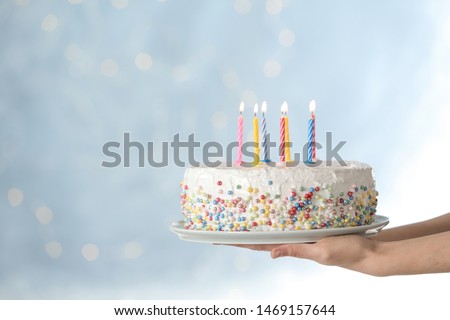 Woman holding birthday cake with burning candles against blurred background, closeup. Space for text
