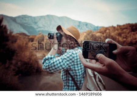 Portrait: Handsome man traveler in a hat and with a backpack on a background of mountains and a lake takes a photo on a professional camera. Traveling blogger traveled