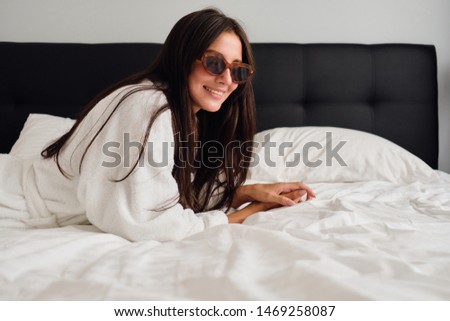 Young beautiful smiling woman with dark hair in white bathrobe and sunglasses happily looking in camera lying on big bed in modern hotel