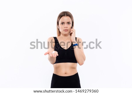 Astonished young sporty blond woman in a black sportswear with wireless ear buds speaking on the phone standing over white background.