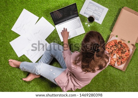 Woman having lunch with pizza and glass of wine working  on the grass using laptop with graphics and charts printed on the paper.