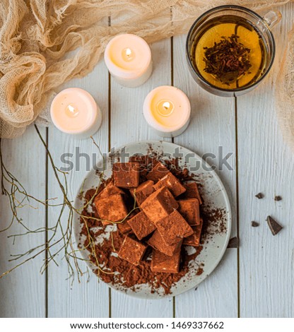 handmade souffle cubes covered with cocoa powder lie on a plate on a light wooden table, hot tea in a glass mug and next to it three white burning candles, pieces of chocolate and a sprig.