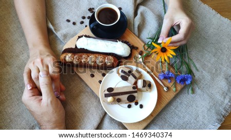 Top view of female and male hands holding each other with summer flowers cornflower and rutbeckia near a cup of black coffee and served eclairs, the concept of a pleasant breakfast, selective focus