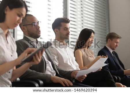Multiracial diverse applicants sit in queue read wait for company work interview, concentrated multiethnic people in row hold papers and gadgets prepare for job talk hiring. Employment concept