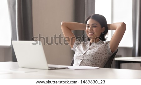 Smiling young woman worker sit in chair hands over head take break from work thinking or dreaming, happy female employee stretch at workplace look at laptop screen watching video online Royalty-Free Stock Photo #1469630123