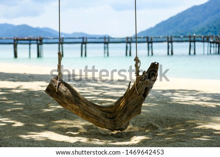 Wooden swing on the beach. The background is a white sand beach and a wooden bridge that extends into the sea and there are mountains in the distance.