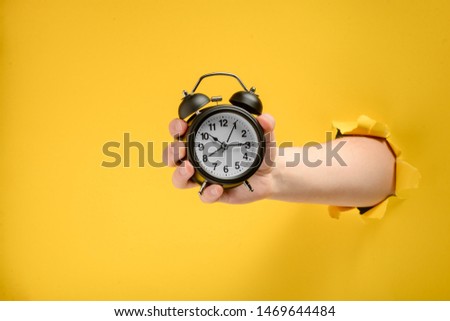 Hand holding a vintage black alarm clock through torn yellow paper background. Wake up on time.