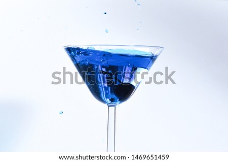 Cocktail with blue liquid in glass. Glass with blue water pouring with liquid with splashes and drops. Martini glass filling with alcohol with splashes on white background. Refreshing drink concept.  