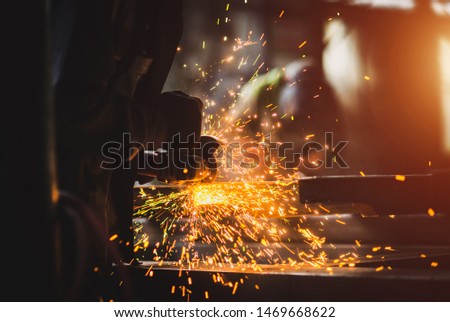 A man grinds metal. In the background a welder welds a part.