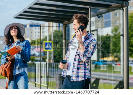 Students couple traveler waiting for tram on stop.. Handsome guy talking phone while elegant woman reading book conspectus on bus station with cityscape on background.