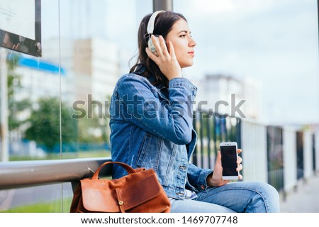 Excited brunette woman wearing blue denim jacket with headphones relaxing, listening music while waiting at tram stop in city.City public transport infrastructure, citizen and tourists transportation.
