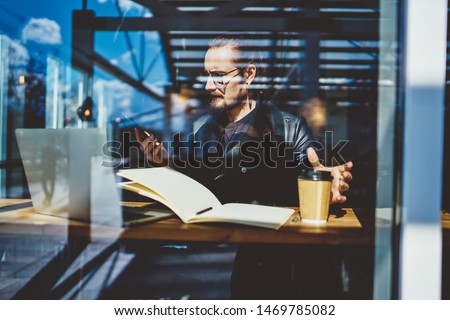 Serious skilled graphic designer working remotely in coffee shop sitting at table with sketchbook and laptop computer while reading received email on cellular phone connected to 4g wireless