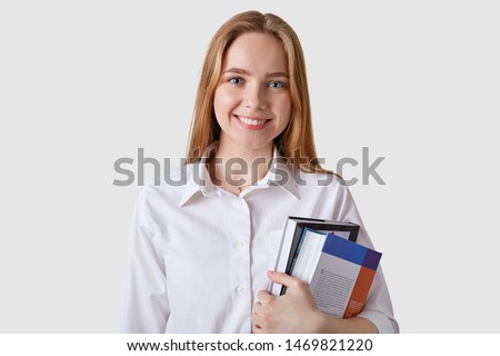 Horizontal shot of college student with broad smile and happy facial expression, glad to hear good news from groupmates, holding folders with papers, isolated over white studio wall. Education concept