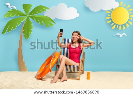 Positive European woman relaxes in deck chair, makes selfie on modern cell phone, shows peace sign, shares pics with friend in social networks, uses sunscreen lotion for good sunbathing. Summer time