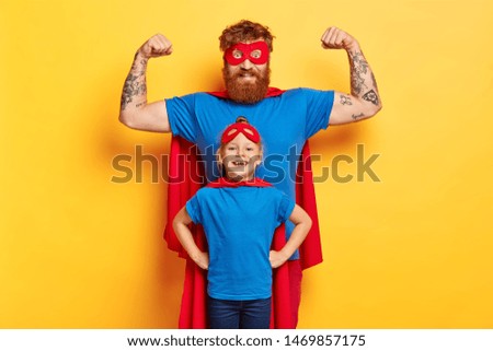 Family fun concept. Joyful strong father raises arms and shows biceps, ready to defend his daughter, stands back, imagine they are superheroes, wears costumes, isolated on yellow background. Royalty-Free Stock Photo #1469857175
