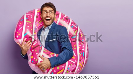 Delighted joyous businessman wears formal clothing, holds inflatable circle, smiles happily, looks with dreamy expression on right side, imagines future vacation at sea, poses indoor over violet wall