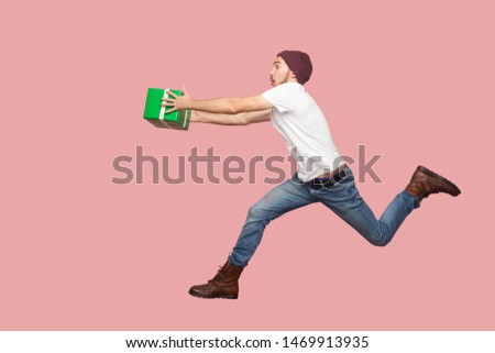 Side view of portrait of crazy bearded young hipster man in white shirt and casual hat jumping, running and hurry up with delivery green present. Indoor, isolated, studio shot, pink background