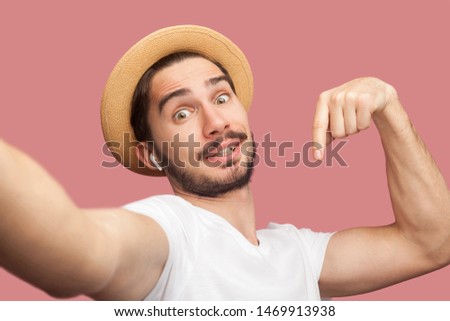 Close up portrait of funny handsome bearded young hipster blogger in white shirt and casual hat posing and making selfie on phone,point finger down. Indoor,isolated, studio shot, pink background