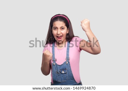 Portrait of rejoicing winner brunette young girl in casual pink t-shirt and blue denim overalls standing, screaming and celebrating her victory. indoor studio shot, isolated on light gray background.
