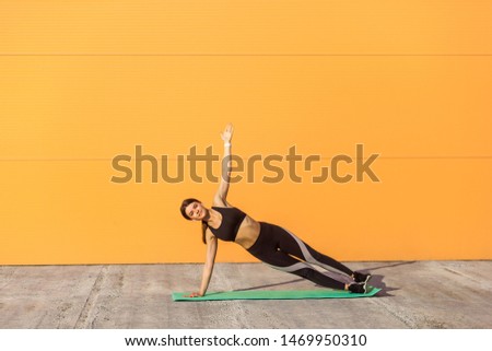 Young sporty attractive woman practicing yoga, doing vasisthasana exercise, side plank pose, working out on green mat, wearing black sportswear, Outdoor, orange background, sport and healthy concept