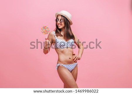 Girl eating Lollipop in swimsuit sunglasses and hat on pink background