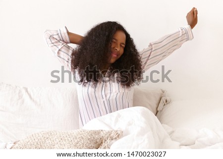 Sleepy pretty girl with brown skin and voluminous hairdo stretching arms, keeping eyes closed, feeling lazy to get up early in the morning. Portrait of stylish cute young African woman in bed Royalty-Free Stock Photo #1470023027