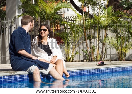 A man and a woman are relaxing together beside a pool.  They are looking at each other and dipping their legs in the water.  Horizontally framed photo.