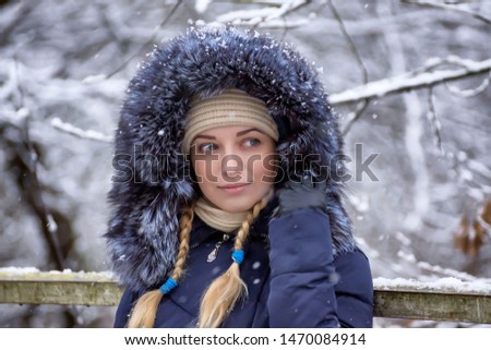 A cute, young, beautiful girl in a blue warm coat with a hood decorated with fur walks in a snowy, winter park.