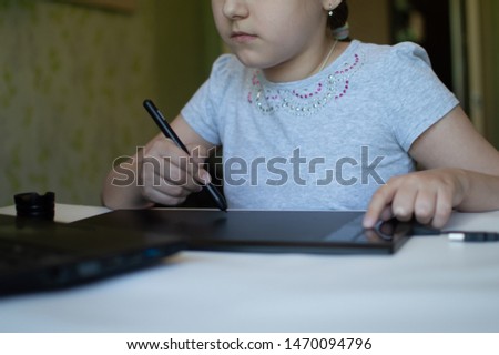 child girl draws on a graphic tablet while sitting at a table with a laptop