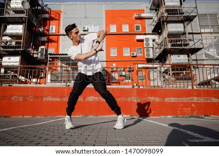 Young guy dressed in jeans and t-shirt is dancing modern dance in the street on the background of urban buildings in the warm day