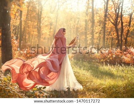 bright summer photo of mysterious beauty in morning forest, lady in shiny white dress and peach pink cloak with long train and hood, back to camera and turned face, girl with dark hair and barn owl Royalty-Free Stock Photo #1470117272