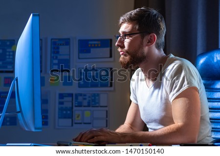 A man with a beard working at the computer. Freelancer develops new programs for mobile devices. Man enthusiastically working at the computer. The designer executes the project.