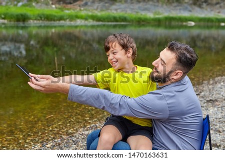 Young bearded caucasian man is sitting near river with his son on his knees and they are taking photos and smiling.