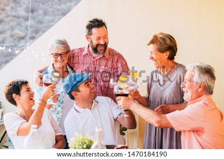 Group of different ages and generations have fun together toasting and clinking with cocktails laughing a lot together - cheerful people with glasses during celebration at home in outdoor terrace 