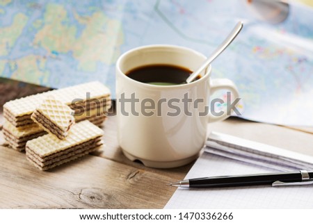closeup of pencil lying on notebook in front of white coffee mug with black coffee next to some waffle bisquits on wooden surface in front of  big map