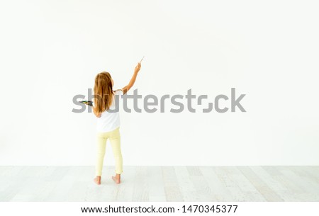 Little girl painting on white wall indoors. view from the back
