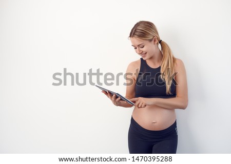 Happy young pregnant woman with bare baby bump or abdomen standing reading on a tablet pc with a pleased smile over a white studio background with copy space